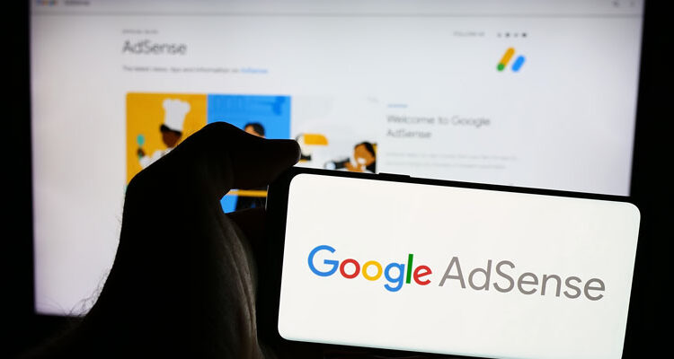 A person holds a smartphone displaying the Google AdSense logo, with the official AdSense website visible blurred in the background on a computer screen.